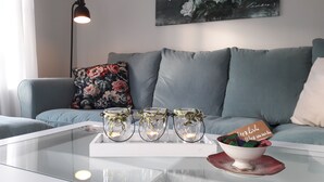 Una sala de estar con una mesa de centro de vidrio, un cojín floral y una bandeja con velas y un cuenco de bombones.