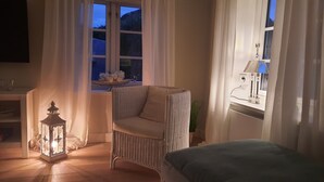 Una habitación acogedora con una silla de mimbre, una mesita con un cuenco de flores, un farol encendido y una cama.
