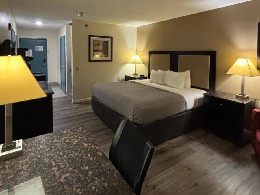 A hotel room with a bed, two bedside lamps, a desk with a chair, a red couch, and a door leading to a hallway.