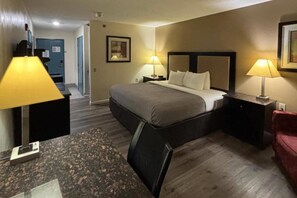 A hotel room with a bed, two bedside lamps, a desk with a chair, a red couch, and a door leading to a hallway.