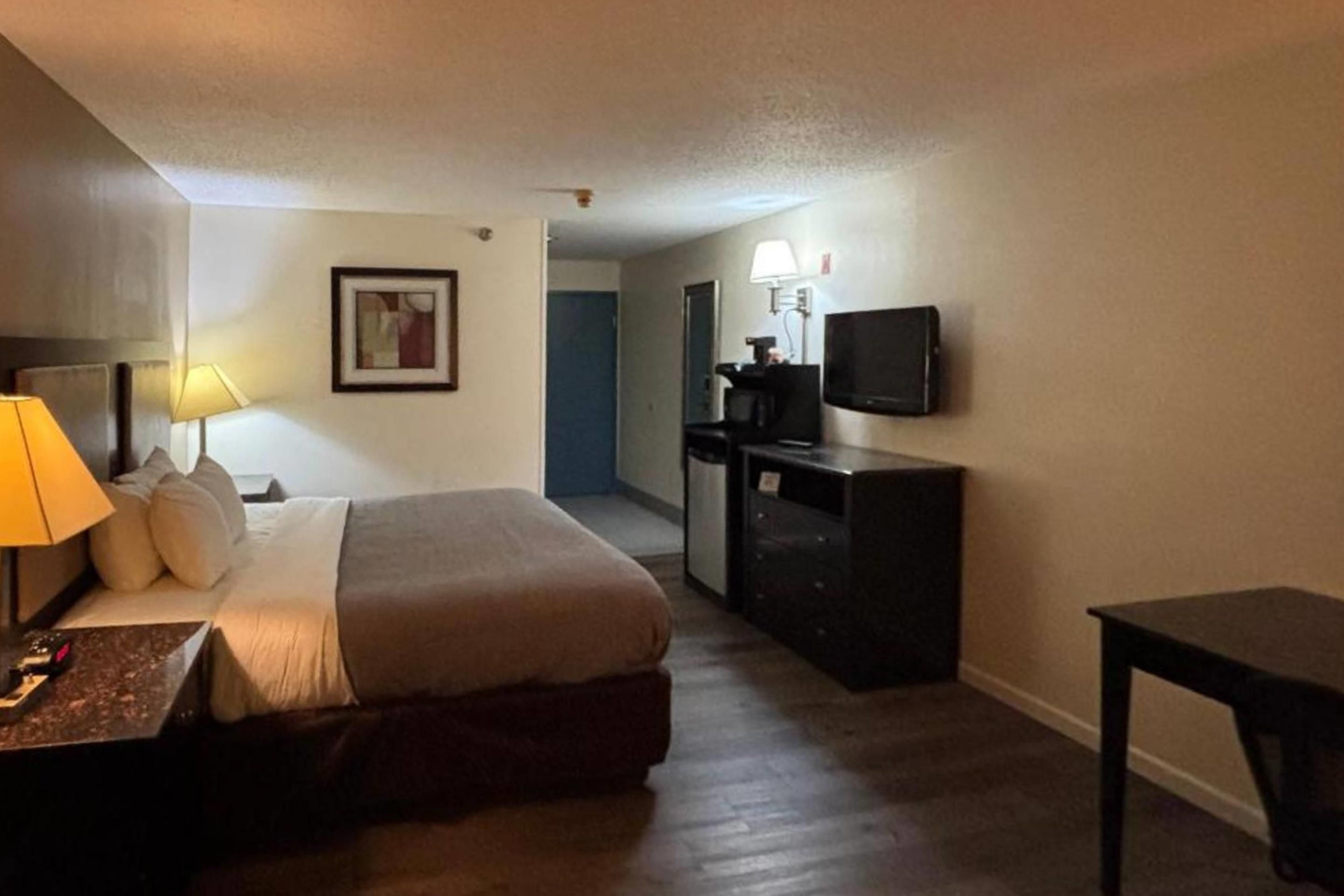 A hotel room with a bed, a desk with a lamp, a dresser, a television, and a door leading to another room.
