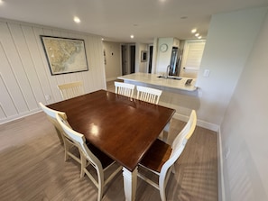 Un coin repas avec une table en bois et des chaises blanches, une cuisine équipée d’un évier et d’un réfrigérateur, et une carte accrochée au mur.