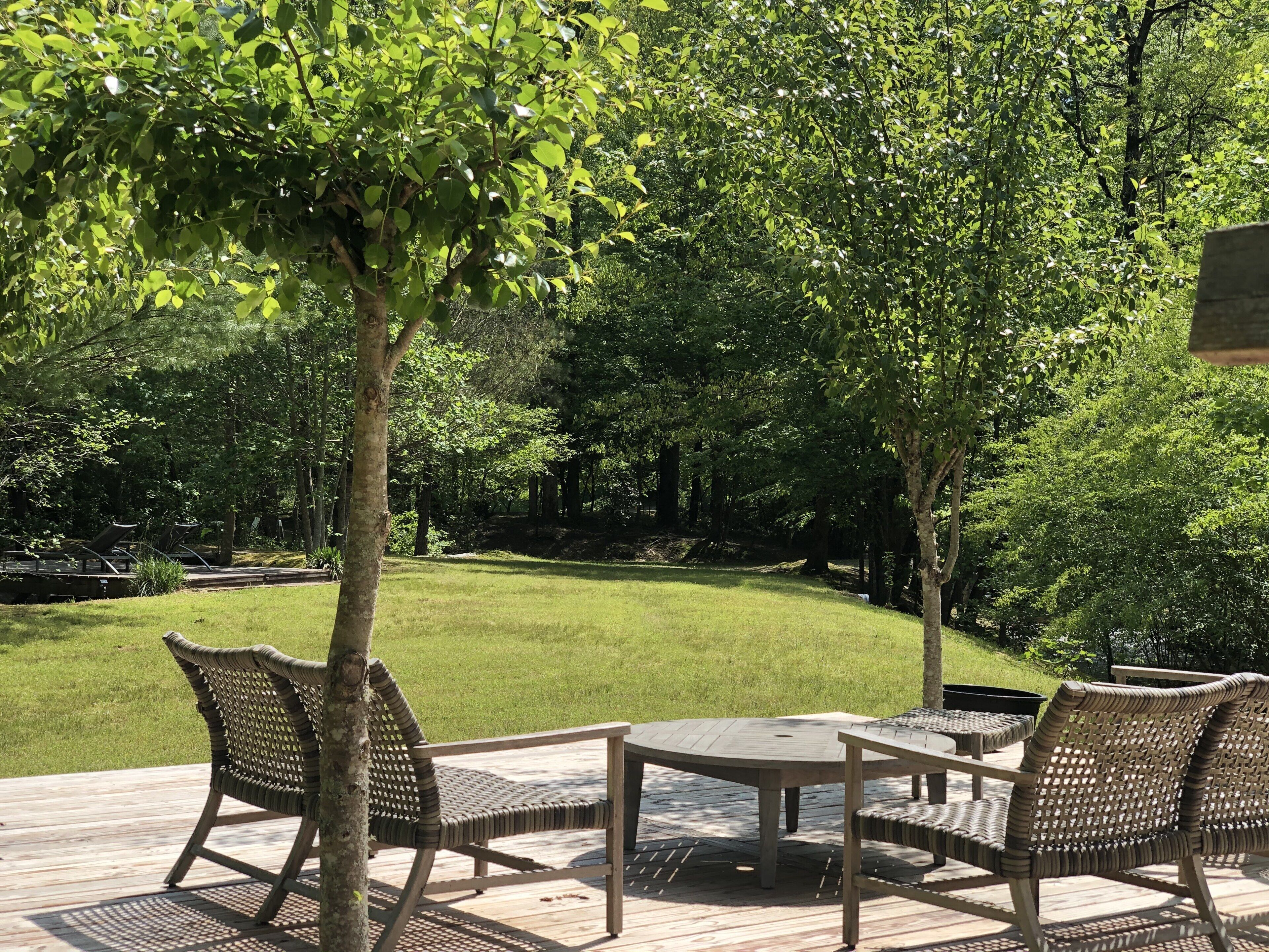 An outdoor deck with wicker furniture and a table, surrounded by lush greenery and trees.