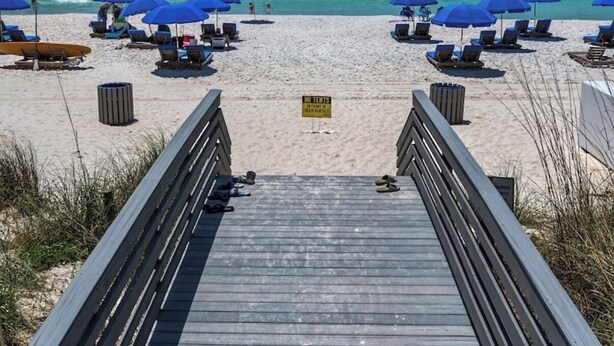 En strand med en træpromenade, der fører til vandkanten, med blå parasoller og liggestole.