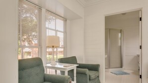 A room with two green upholstered chairs, a white table with a lamp, and a view of a shower area through a glass door.