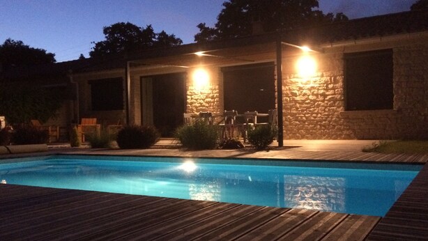 Ein Pool mit Holzterrasse, Steinwänden und Beleuchtung im Freien.