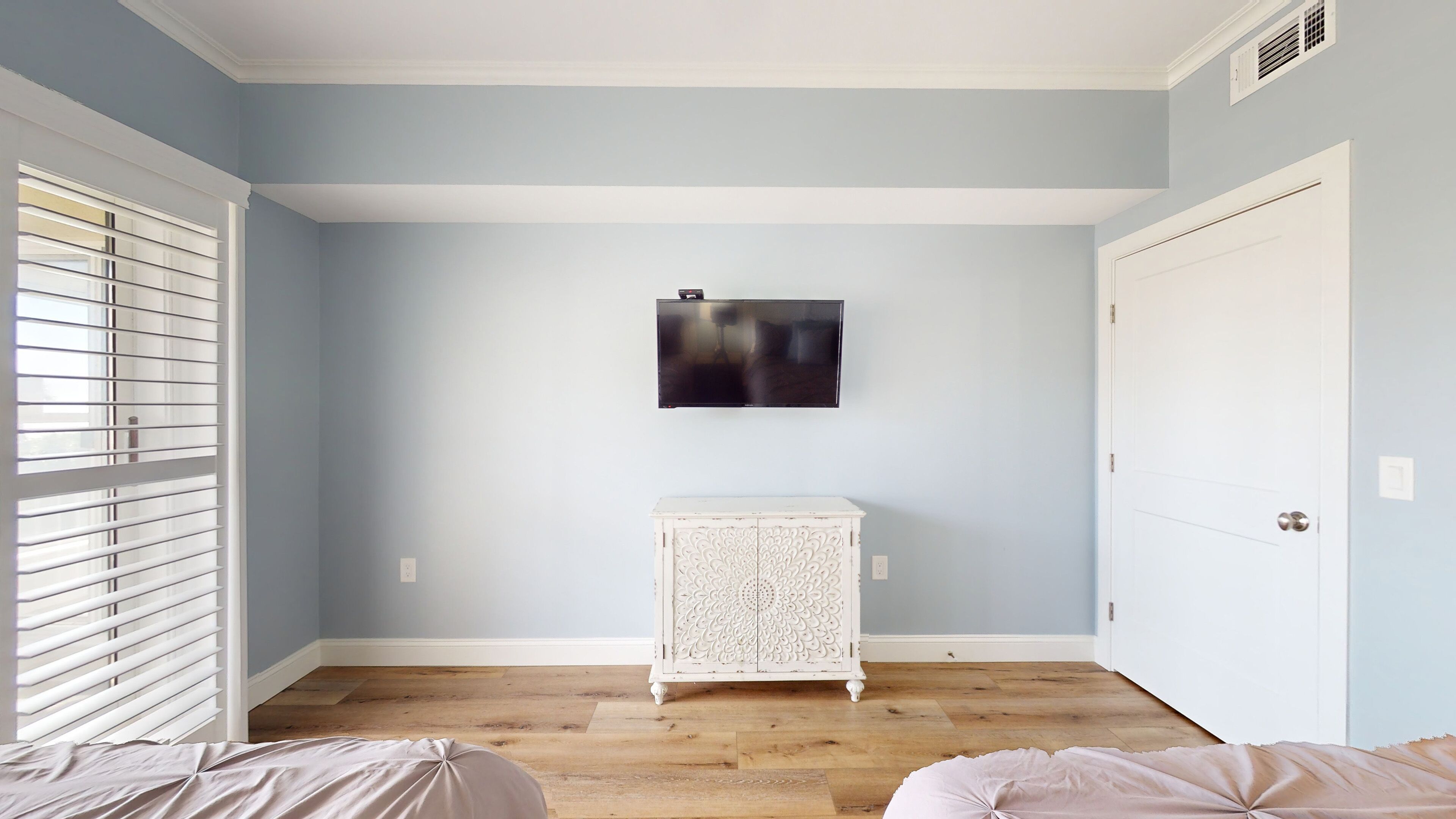 Ein Zimmer mit hellblauen Wänden, Holzboden, einem weißen Schrank, einem an der Wand befestigten Fernseher und einer Tür rechts.