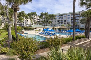 A resort with a swimming pool surrounded by blue lounge chairs and umbrellas, palm trees, and a multi-story building with balconies.