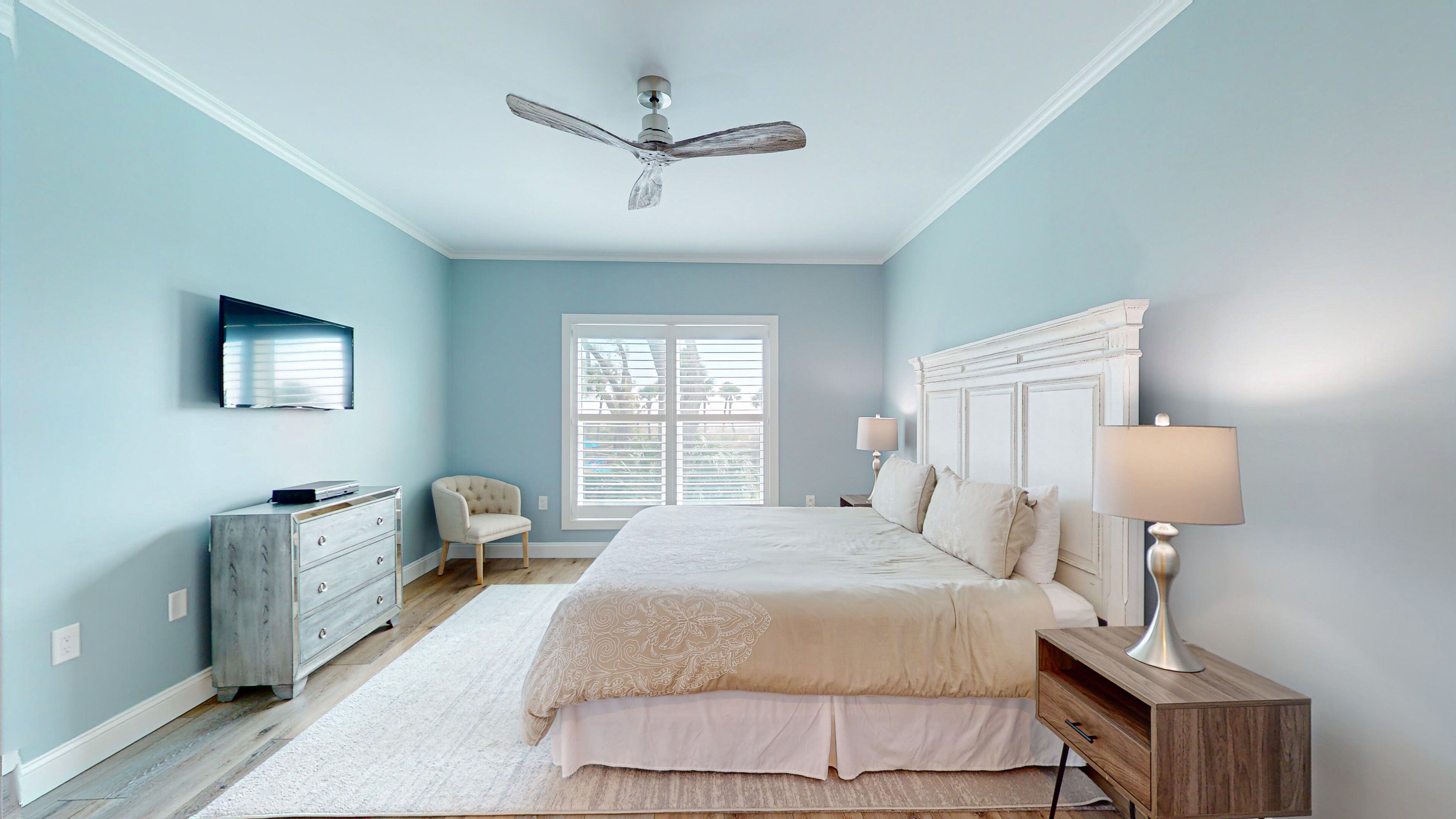 Une chambre à coucher comprenant un lit, une chaise, une commode, une table de chevet, une lampe, un ventilateur de plafond et une fenêtre.