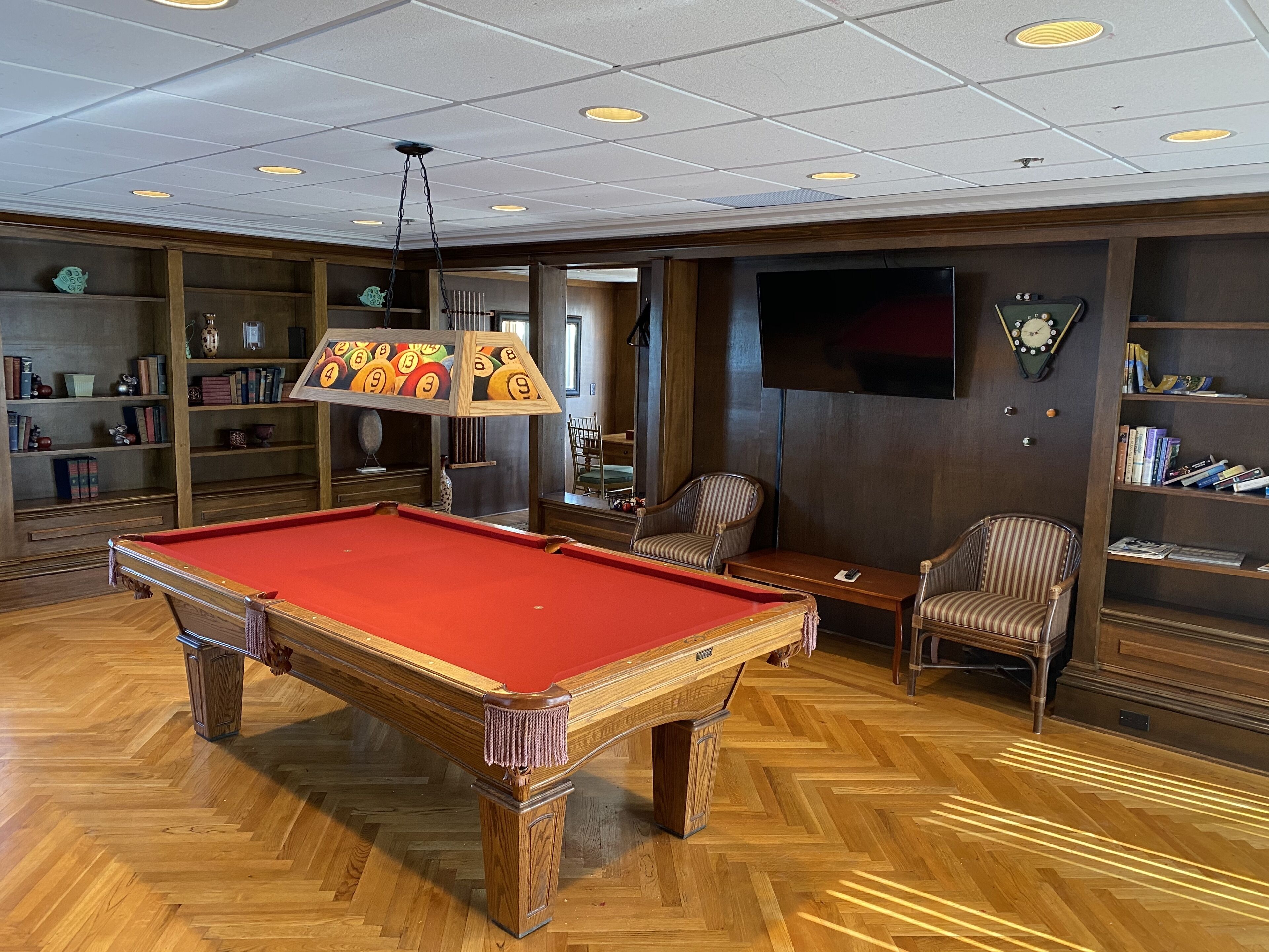 A pool table in a room with wooden flooring, a bookshelf, a television, and a clock on the wall.