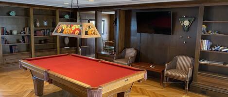 A pool table in a room with wooden flooring, a bookshelf, a television, and a clock on the wall.