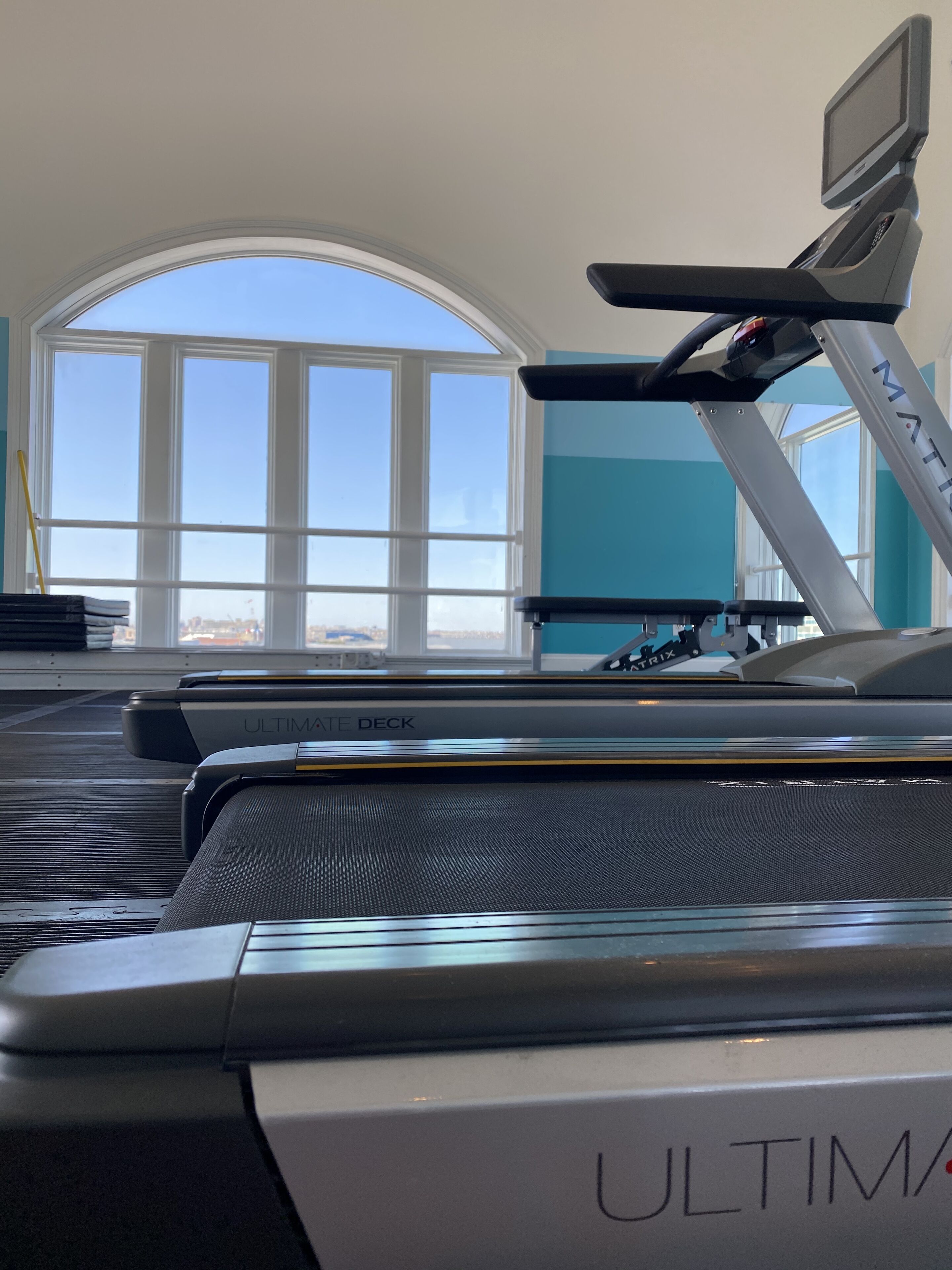 A treadmill with a digital display, positioned in a room with large windows and a view of the cityscape.