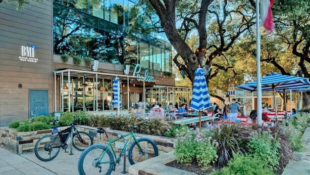 An outdoor seating area with tables and chairs, a bicycle parked nearby, and a building with a glass facade and outdoor seating. - Chic SoCo Studio | Balcony + Walkable Austin (Austin)