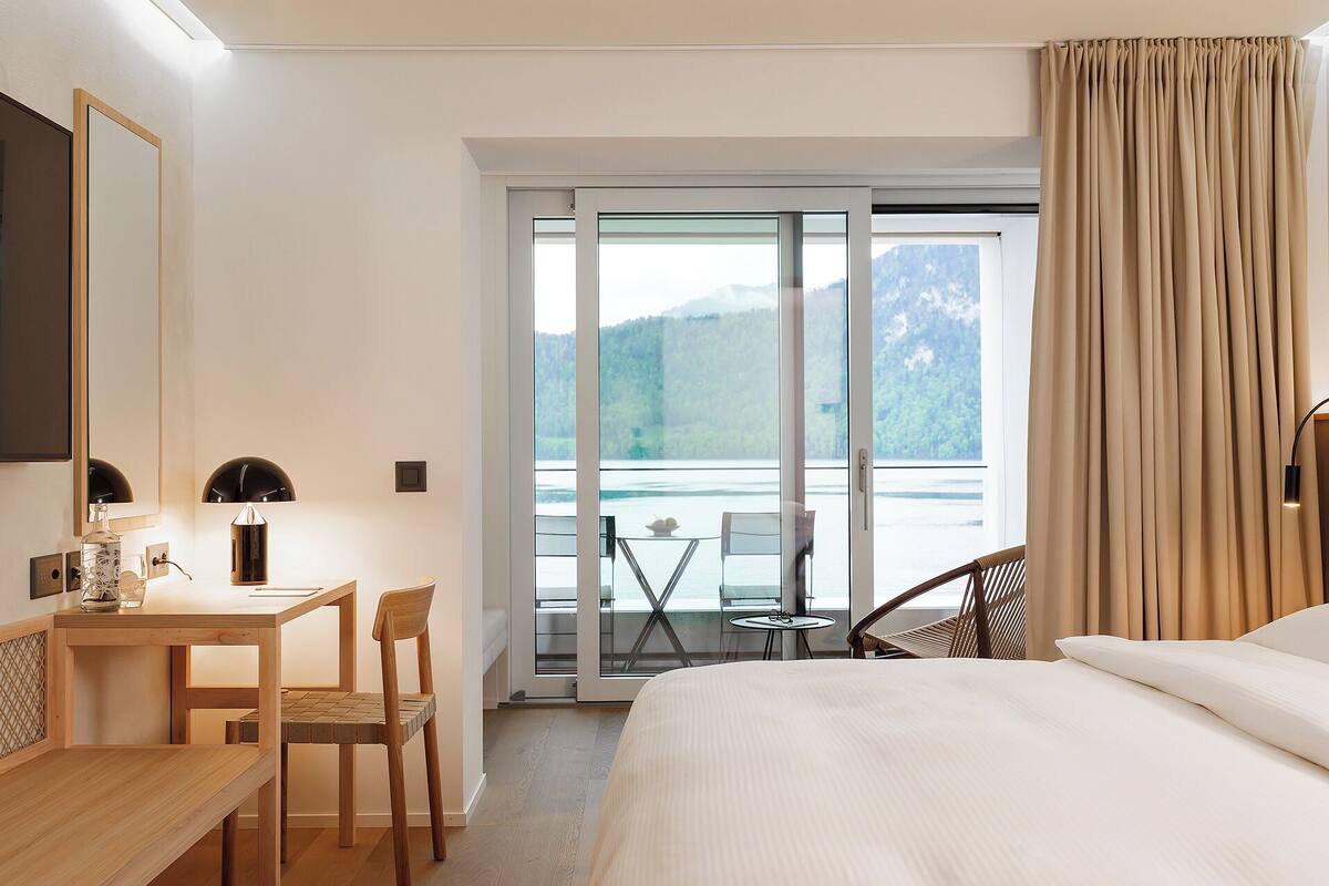 Ein Hotelzimmer mit Bett, Schreibtisch mit Stuhl, einem kleinen Tisch ebenfalls mit Stuhl, einem großen Fenster mit Bergblick und einem Vorhang.