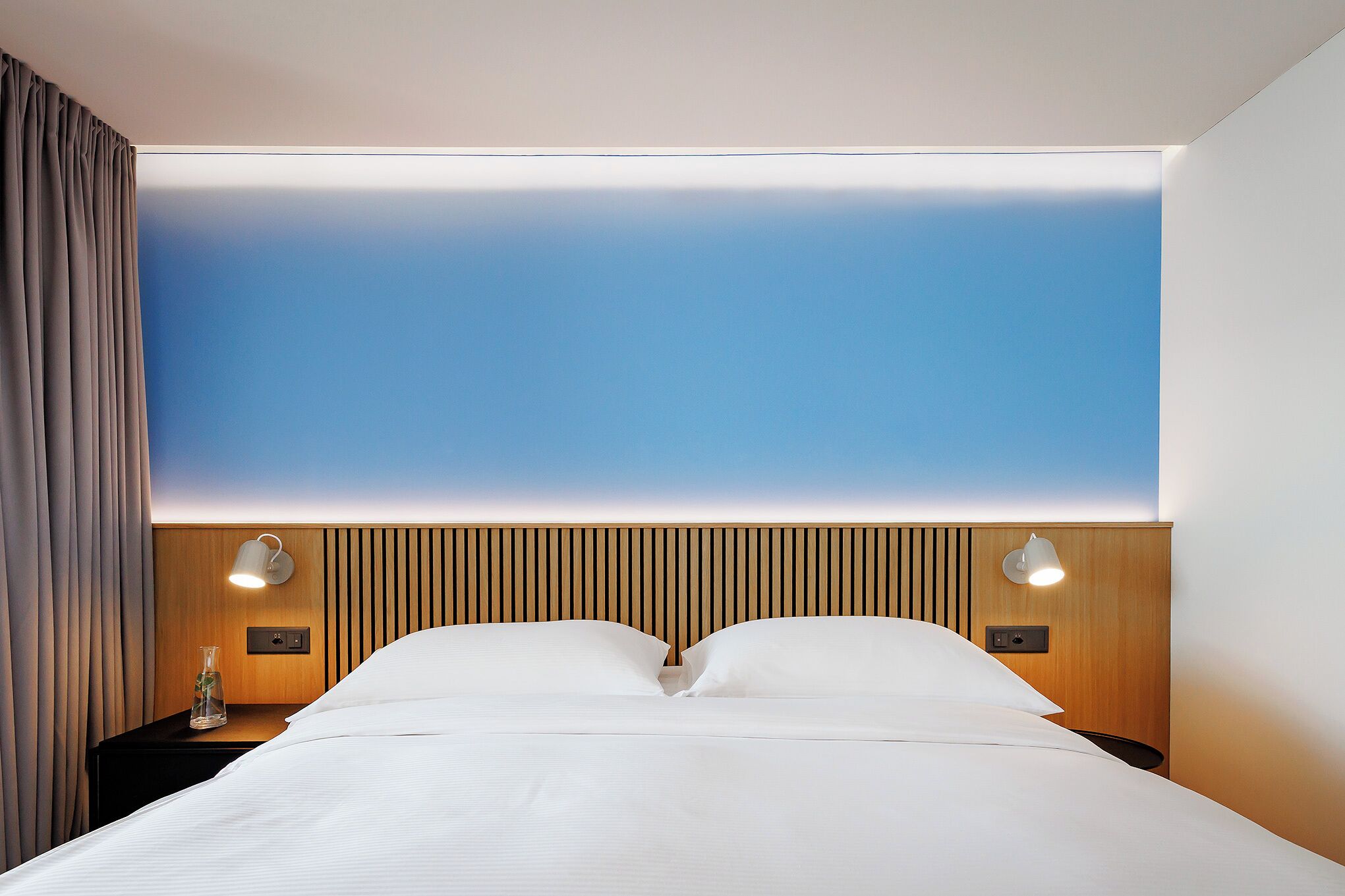 Une chambre d’hôtel moderne avec un grand lit, une tête de lit en bois, deux lampes de chevet et une table de chevet agrémentée d’un vase.