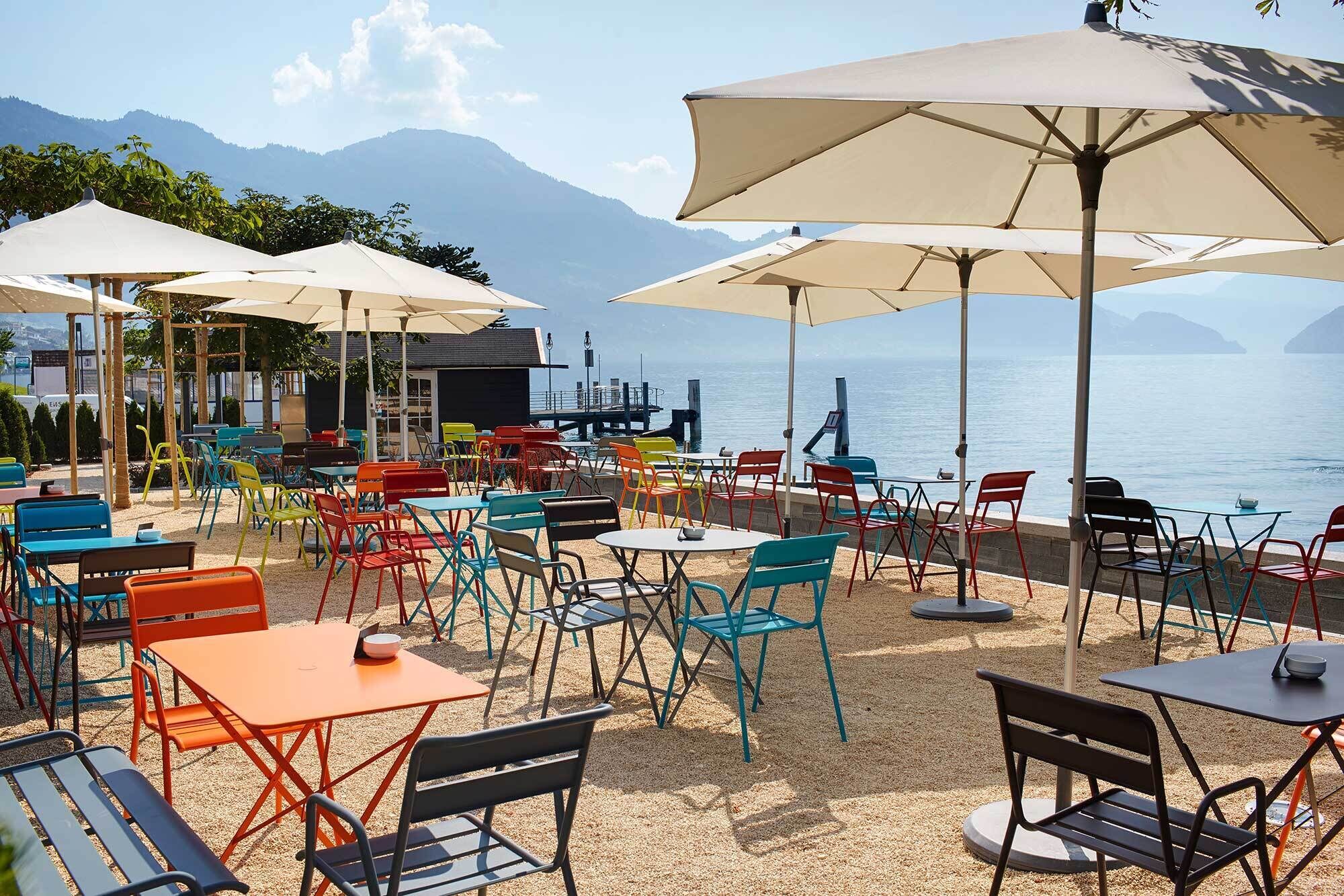 An outdoor seating area with tables and chairs, umbrellas, and a view of the sea and mountains.