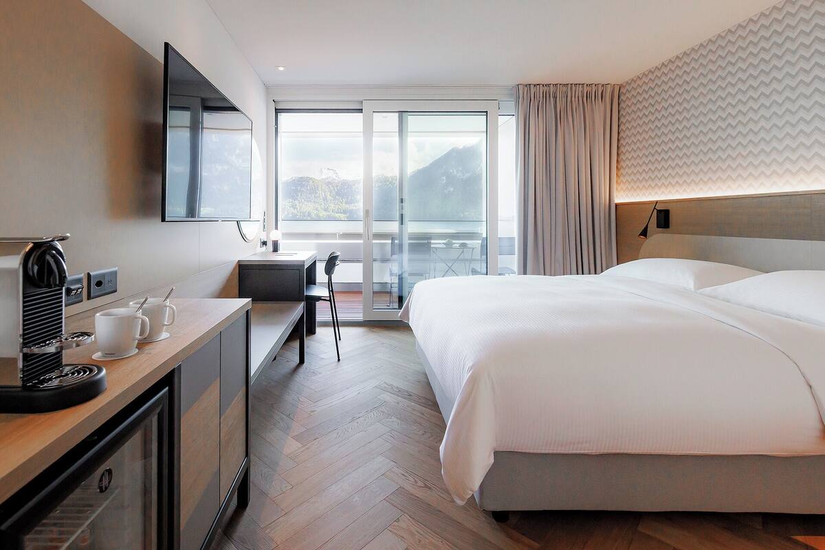 Ein Hotelzimmer mit einem großen Bett, einem Schreibtisch mit Kaffeemaschine, einem Sessel und Blick auf Berge durch das Fenster.