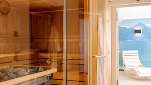 Una sauna con interior de madera, puerta de cristal y vistas a una montaña y al mar a través de una puerta abierta.