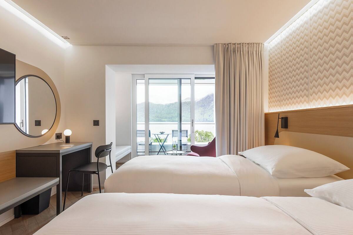 Ein Hotelzimmer mit Bett, Schreibtisch, Sessel und einem großen Spiegel, mit Blick auf einen Balkon inklusive Tisch und Stühlen.
