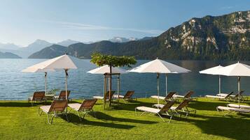 A lakeside terrace with sun loungers and umbrellas, overlooking a mountain range.