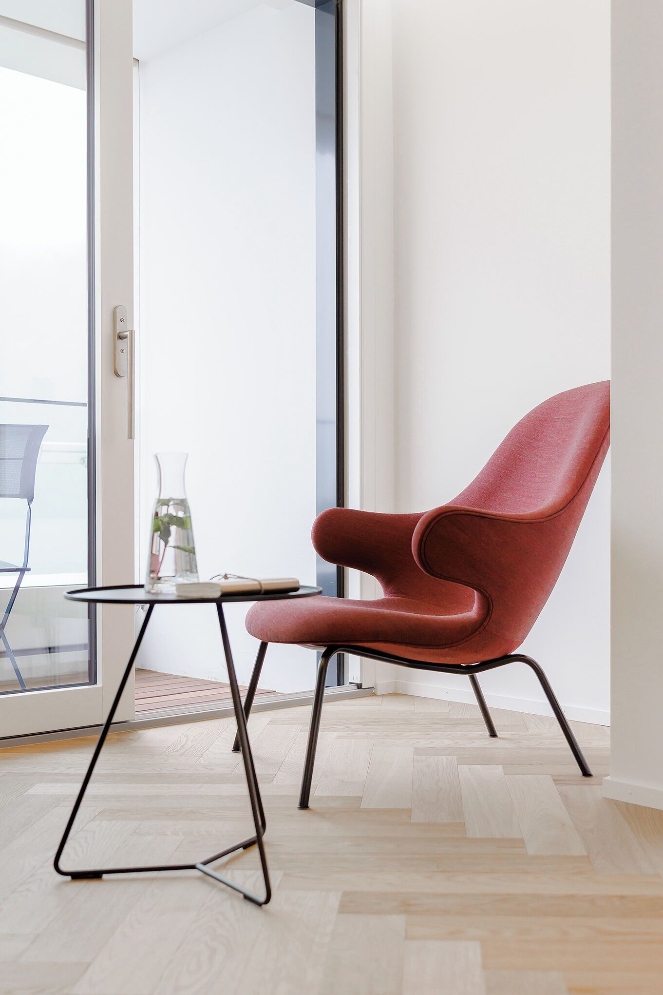 Una habitación moderna con una silla roja, una mesita con un florero y vistas a través de un ventanal.
