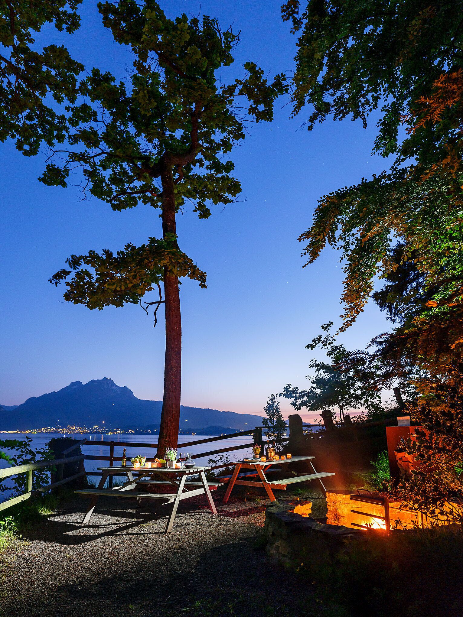 Un espace de pique-nique au bord du lac, avec des bancs en bois et un foyer, entouré d’arbres et offrant une vue sur un lac paisible, avec des montagnes en arrière-plan.