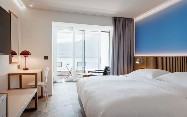 A modern hotel room with a large bed, a desk with a chair, a small table with a chair, a large window with a view of the outdoors, and a wall-mounted lamp. - Campus Hotel Hertenstein (Weggis)