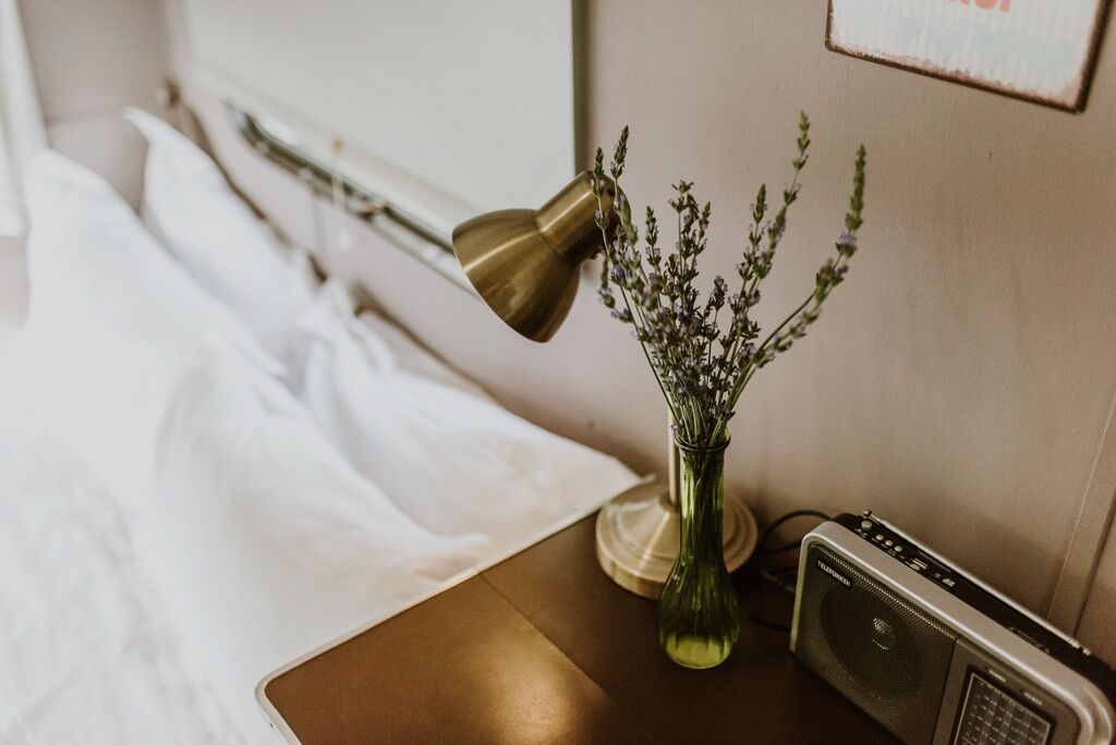 A neatly made bed with white linens, a brass desk lamp, a green vase with lavender flowers, and a vintage radio on a wooden nightstand.