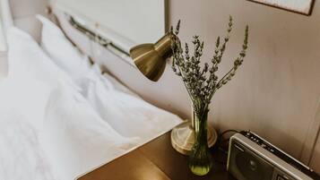 A neatly made bed with white linens, a brass desk lamp, a green vase with lavender flowers, and a vintage radio on a wooden nightstand.
