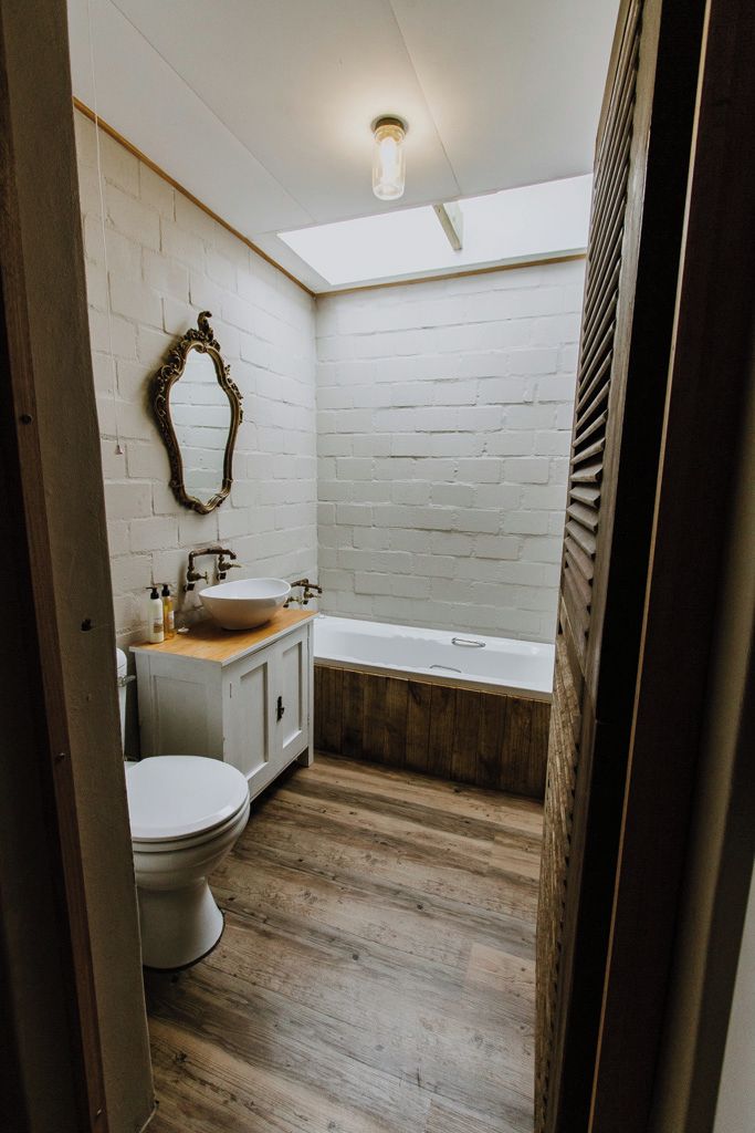 Un baño con pared de ladrillo blanco, suelo de madera, inodoro blanco, tocador de madera, lavamanos blanco, bañera y un tragaluz.
