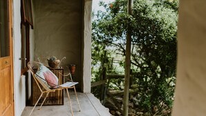 A rustic outdoor seating area with a metal chair and a small table, a wooden door, and a view of a garden with trees and a fence. - The Stables Cottages (Langebaan)