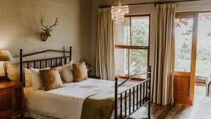 A bedroom with a bed, a nightstand, a chandelier, a window with curtains, and a deer head mounted on the wall. - The Stables Cottages (Langebaan)