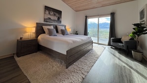 Ein Schlafzimmer mit Holzboden, einem großen Bett mit grauen Bettwaren, einem Nachttisch mit Lampe, einem Sessel und Bergblick durch eine Schiebetür.