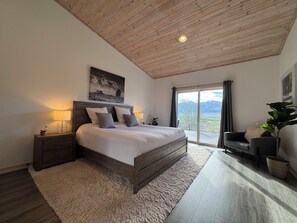A bedroom with a large bed, a nightstand, a lamp, a chair, a potted plant, and a view of mountains through a sliding door.