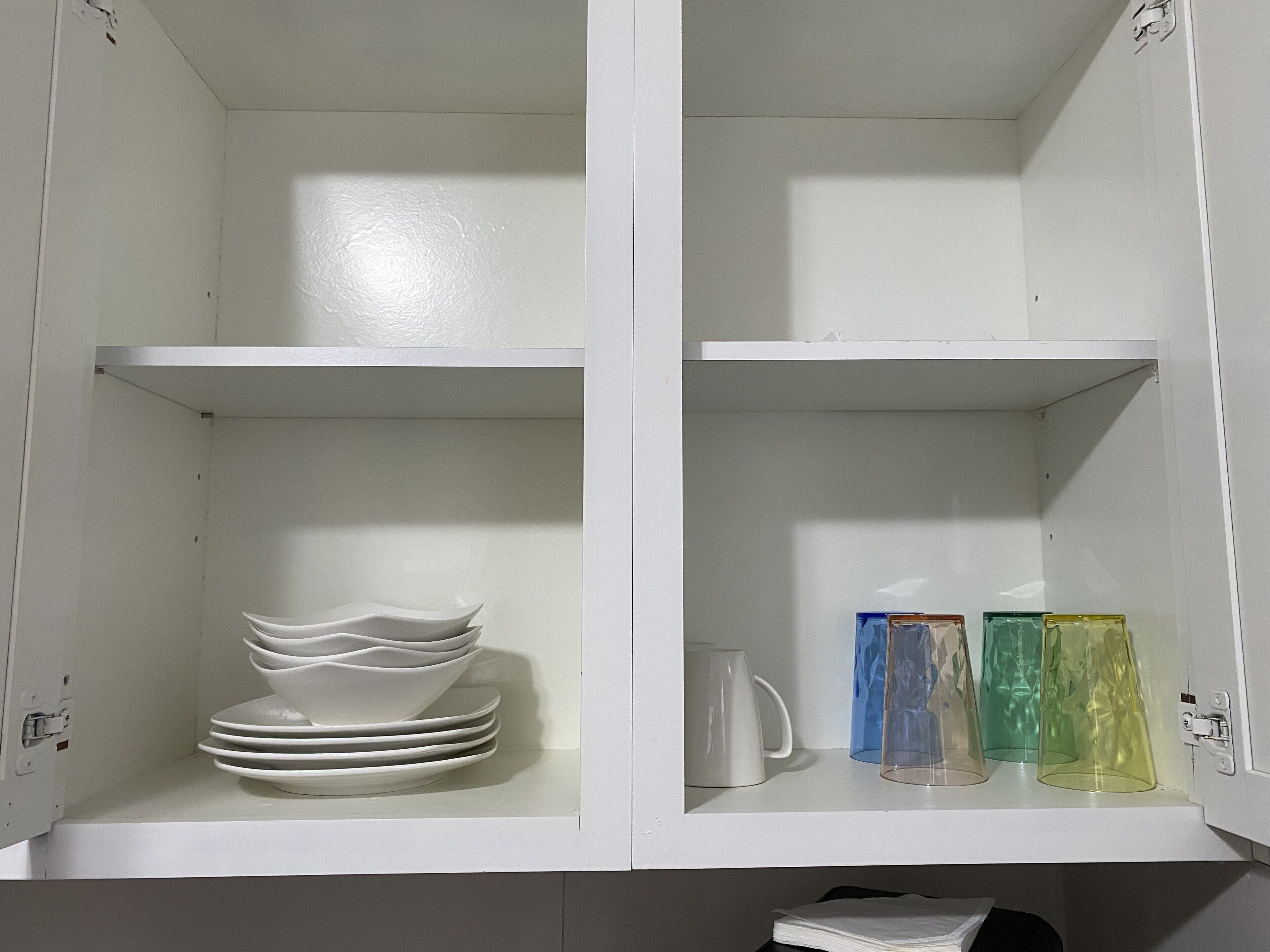 Un meuble blanc doté d’étagères où sont rangées des assiettes blanches, un pichet blanc et des verres transparents de couleur bleue, verte et jaune.