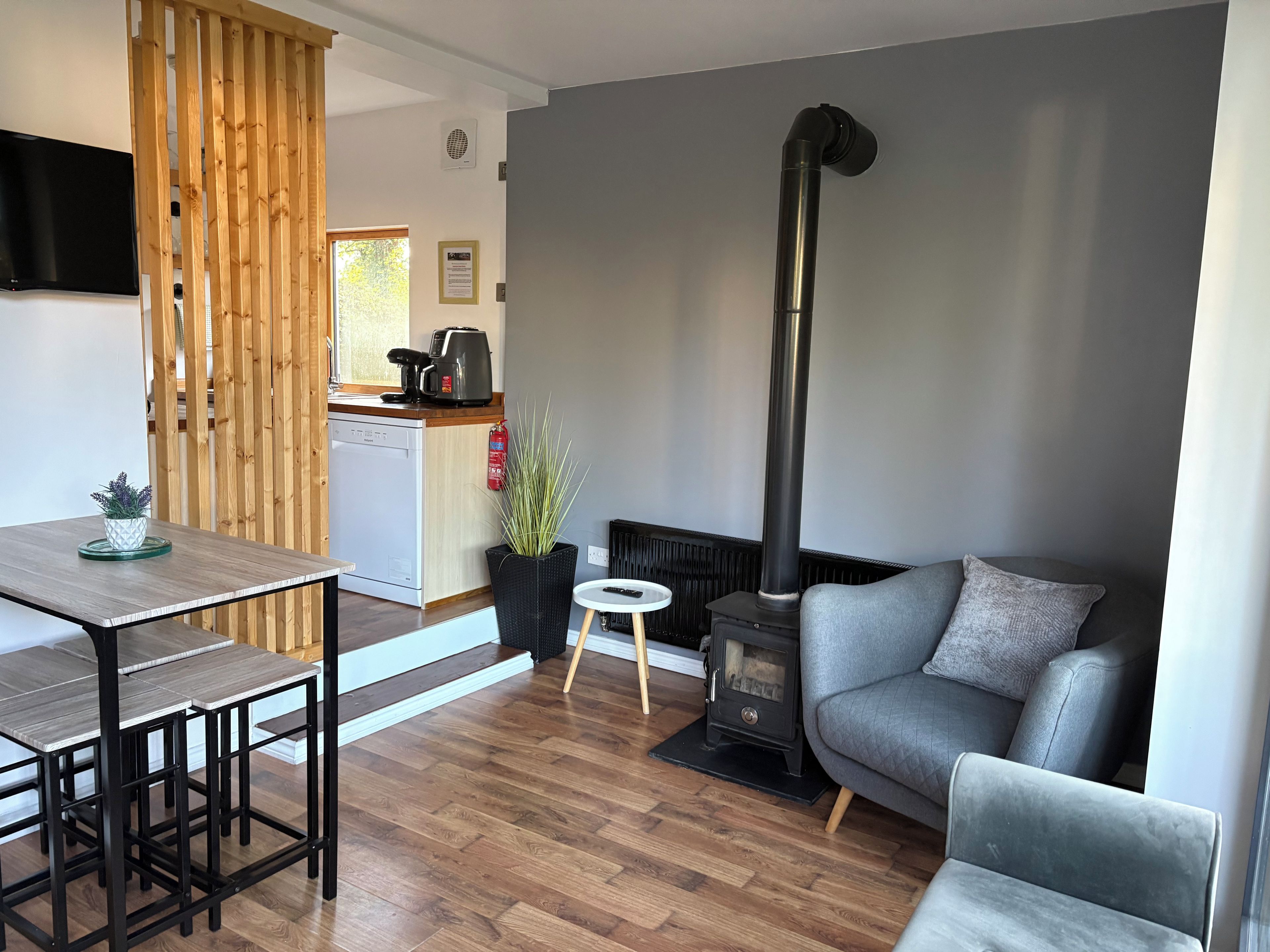 A modern living room with a grey sofa, a dining table with chairs, a kitchen area with a coffee maker, and a black wood-burning stove.