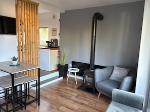 Una sala de estar moderna con un sofá gris, una mesa de comedor con sillas, una zona de cocina con cafetera y un estufa de leña negra.