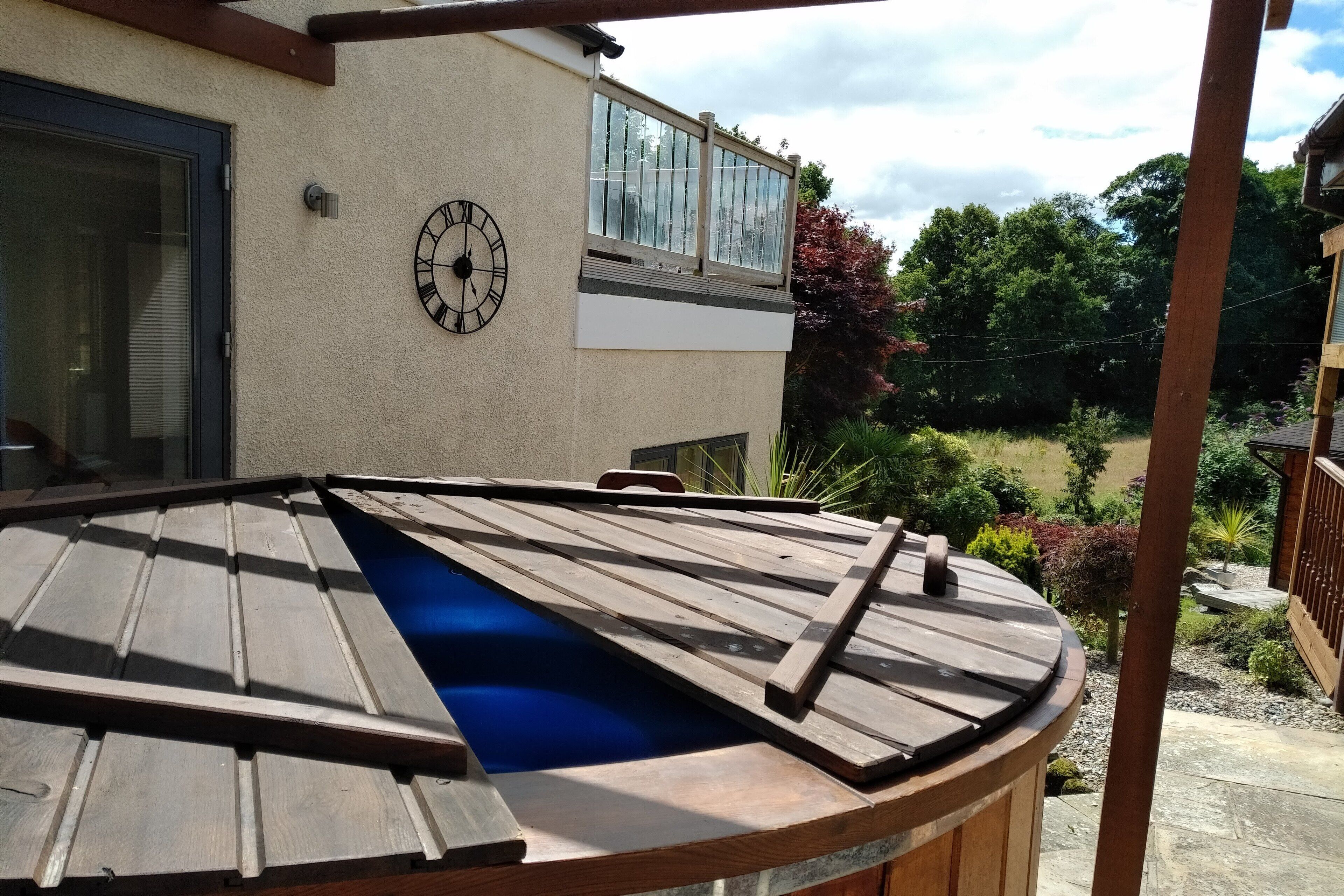 A hot tub with a wooden frame and a blue interior, positioned under a pergola with a wooden roof.