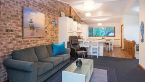 A living room with a brick wall, a gray sofa, a coffee table, and a dining area with a window view. - Fantastic Beach House on Quindalup Strip (Quindalup)