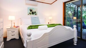 A bedroom with a white bed, two bedside tables with lamps, a green throw blanket, and a window with a view of trees. - Fantastic Beach House on Quindalup Strip (Quindalup)