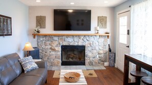 A living room with a stone fireplace, a flat-screen TV mounted above it, a sofa with a decorative pillow, and a wooden coffee table with a woven basket.