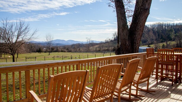 A deck with wooden chairs and tables, overlooking a grassy field and distant mountains.