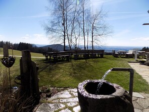 Surrounded by nature, this serene setting features a stone well and picnic tables amidst a grassy area with a view of the surrounding landscape.