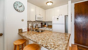A kitchen with granite countertops, white cabinets, a microwave, a refrigerator, and a clock on the wall.