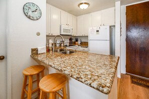 A kitchen with granite countertops, white cabinets, a microwave, a refrigerator, and a clock on the wall.