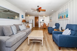 A living room with a grey sofa, blue armchairs, a wooden coffee table, and a dining area with a table and chairs.