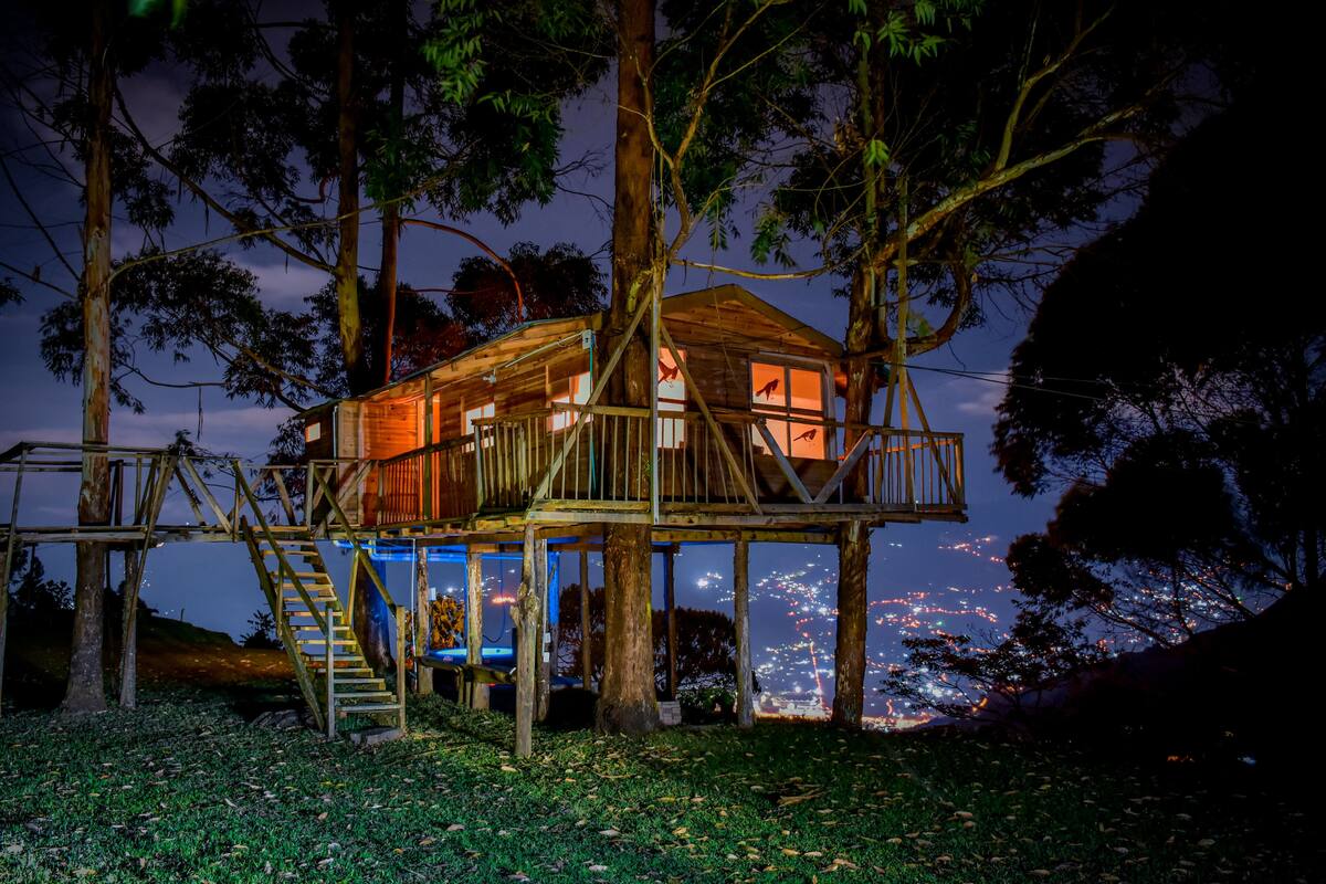 Una casa en el árbol de madera con balcón y escaleras, rodeada de árboles y con vistas a un paisaje urbano nocturno.