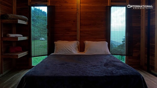 A wooden cabin room with a bed, two windows, and a shelf with towels.