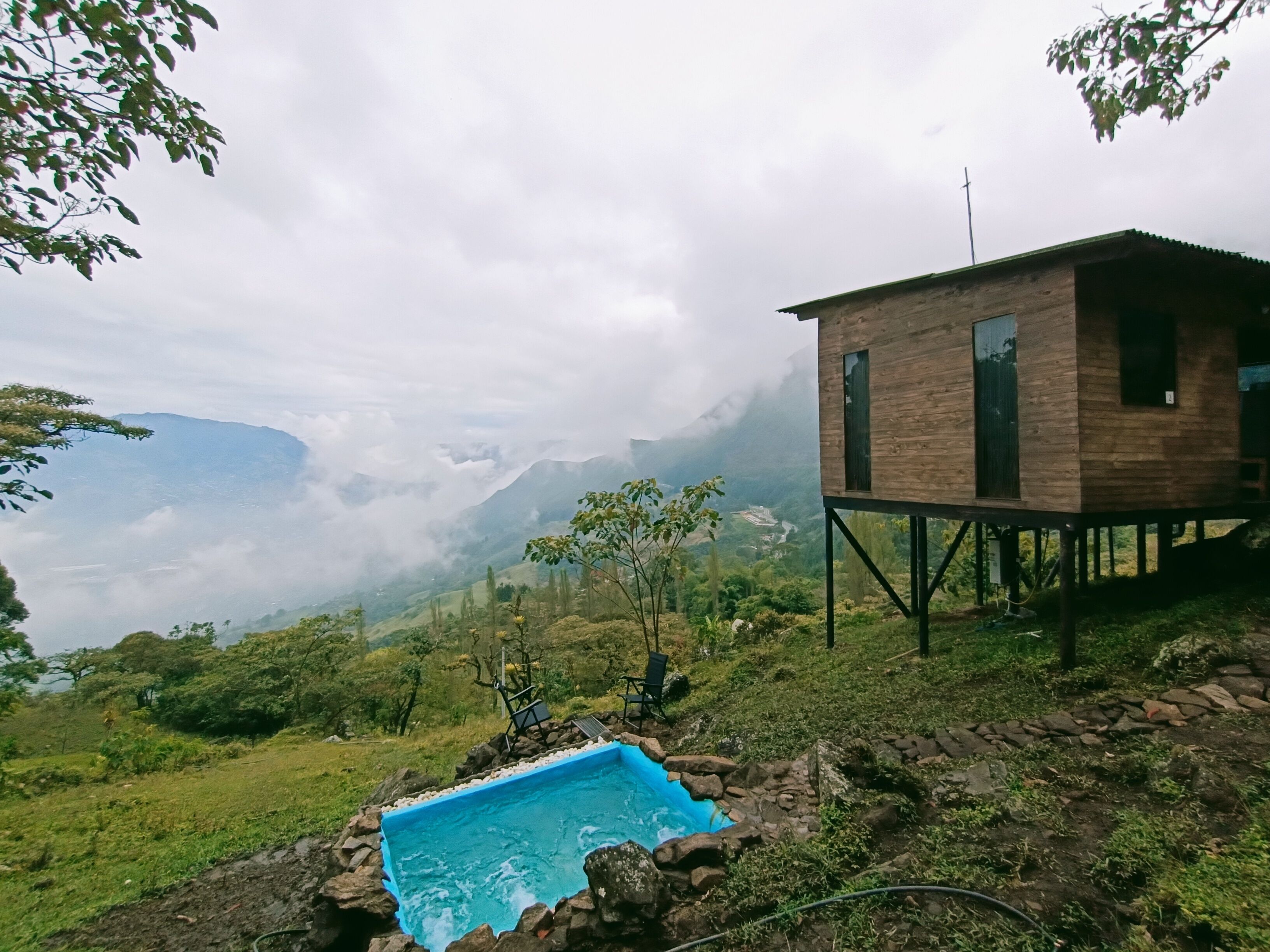 A wooden cabin with a pool on a hillside, surrounded by greenery and mountains.