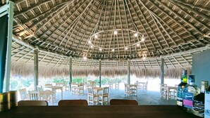 Un área de comedor al aire libre con techo de paja, mesas y sillas de madera, y una barra con botellas.